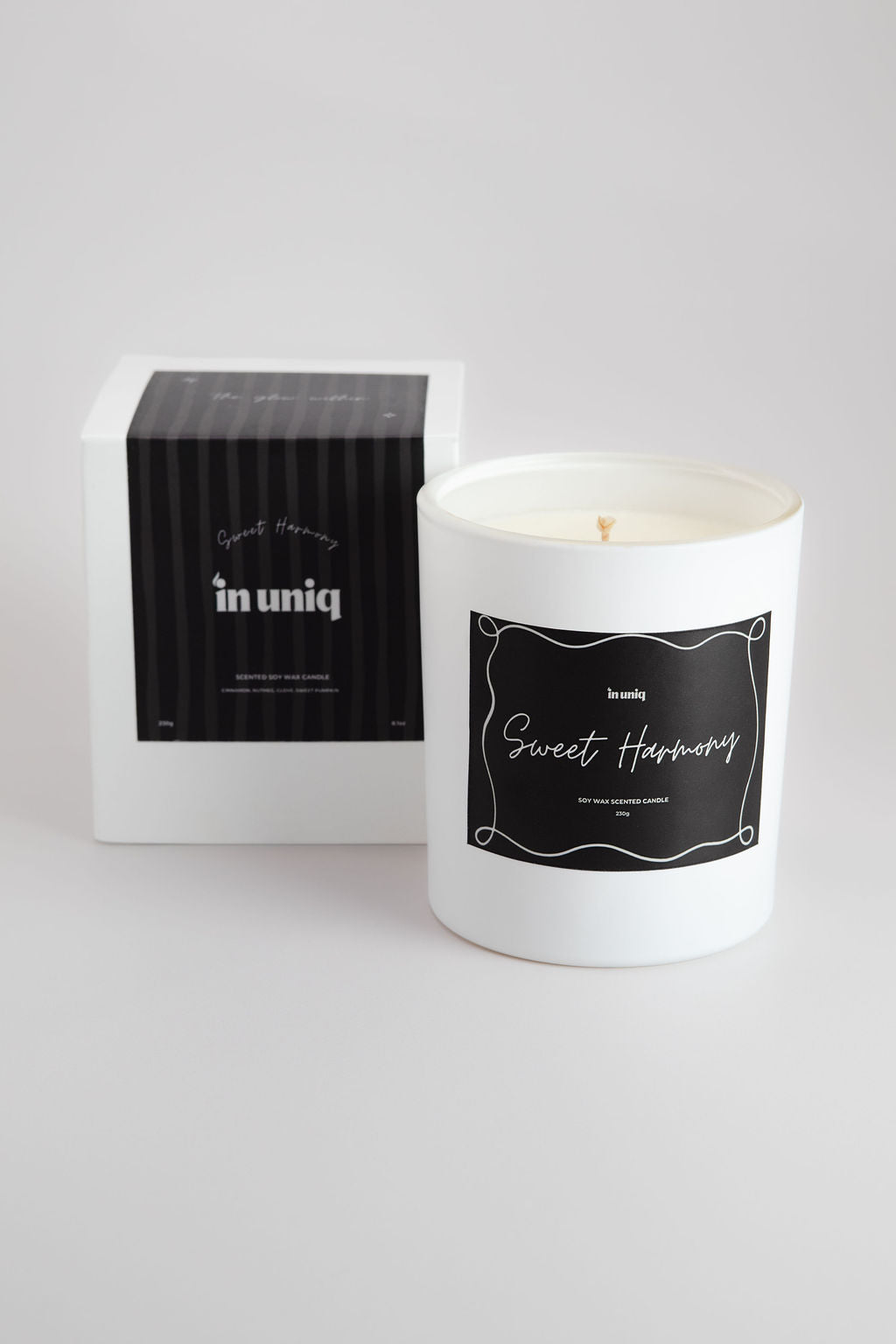 A white glass jar candle with a black label reading 'Sweet Harmony' stands next to its white and black striped packaging box on a light gray background.