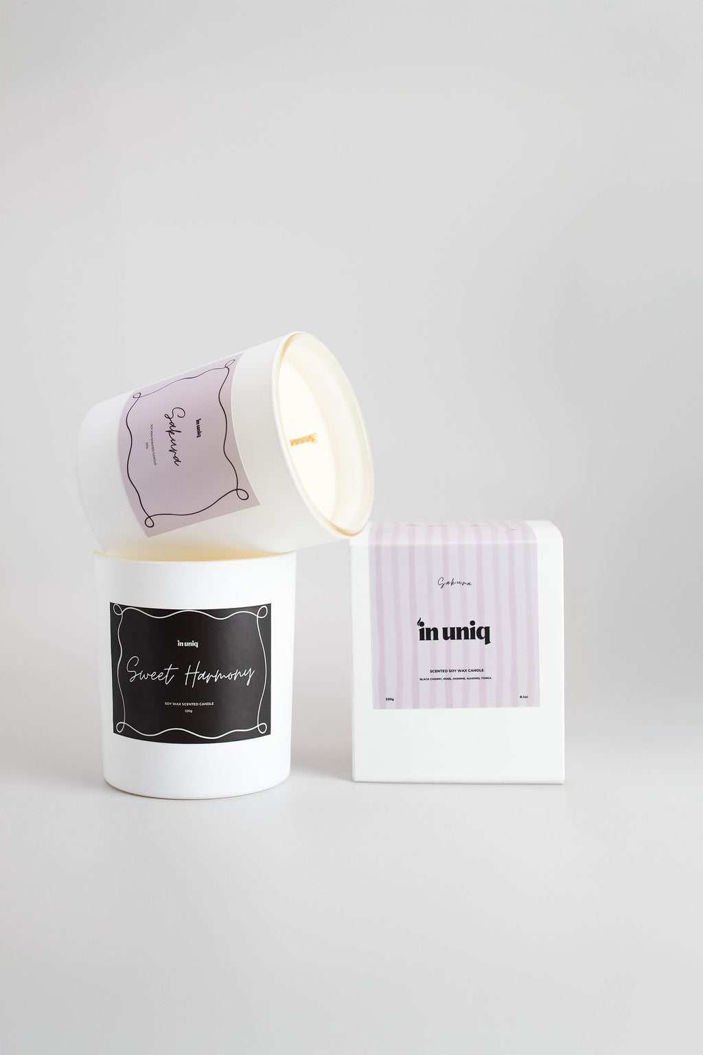 A white jar candle with a dark brown label reading 'Sweet Happiness' sits next to its white box and a second view of the candle showing a pale pink label. The brand name 'in uniq' is visible on the packaging against a light gray background.