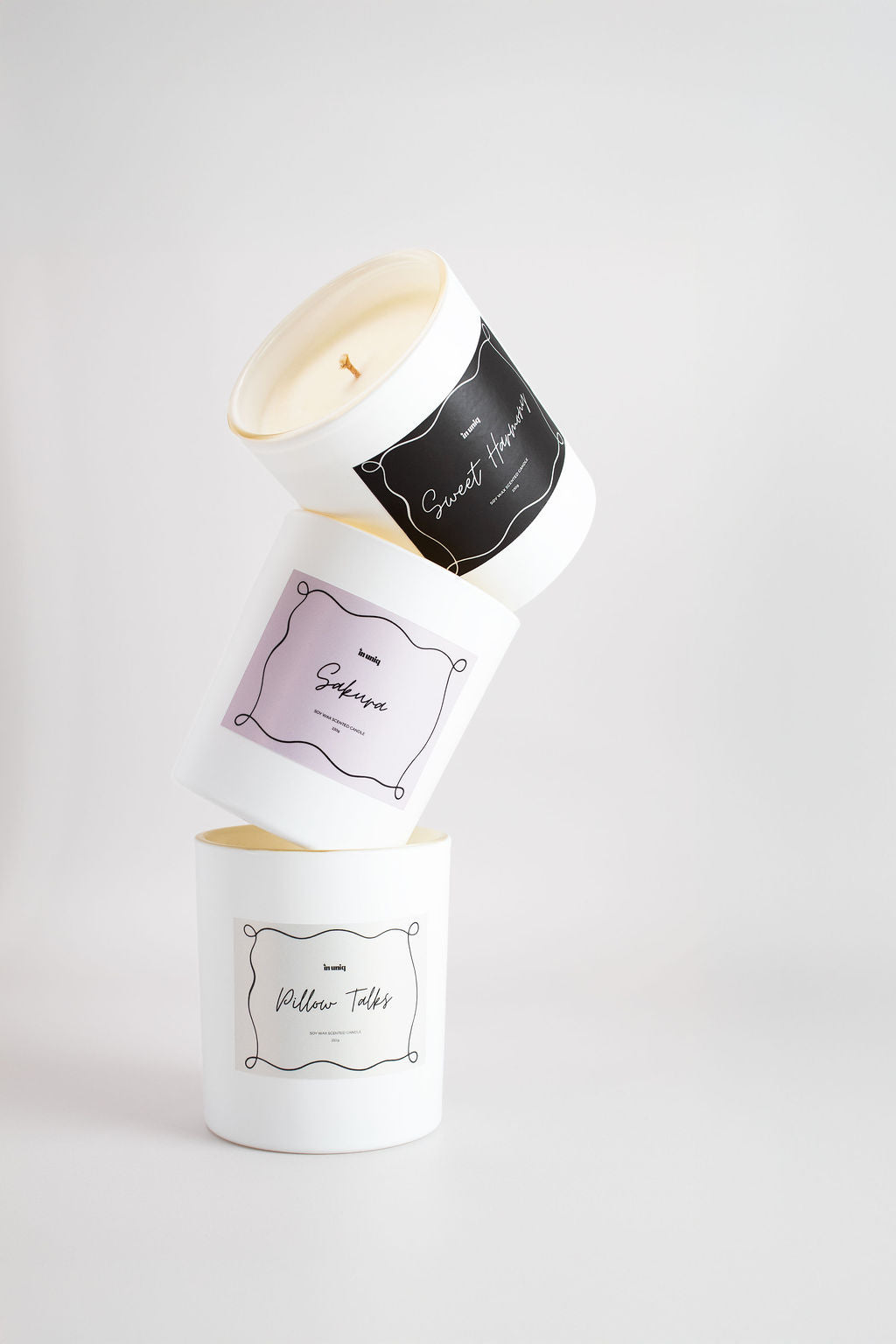 Three white soy candles in cylindrical jars are stacked and balanced in a tower-like arrangement on a light gray studio background. The top candle is tipped on its side, showing a black label with white script, while the middle and bottom candles are upright with pale lavender and black labels respectively.