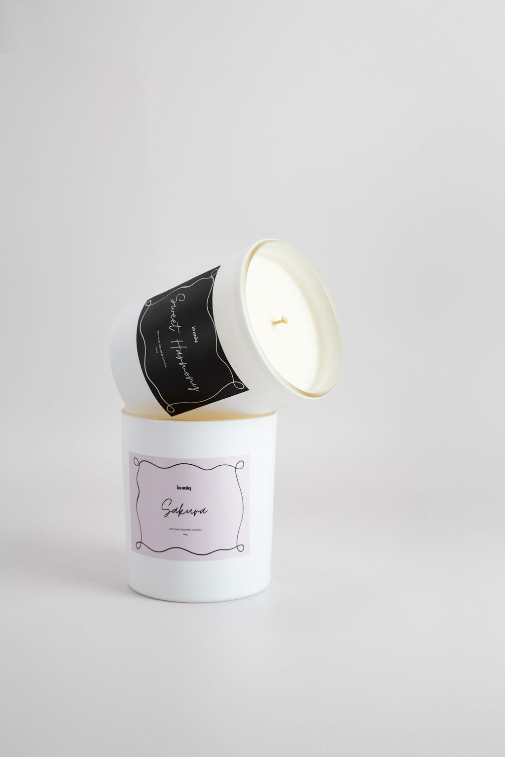 A white soy wax candle jar with a brown label and a second white jar resting on top, photographed on a clean light grey studio style background.